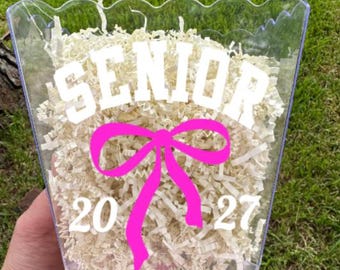 May include: A clear plastic container with a scalloped top edge, filled with light-colored shredded paper filler. The front features white text reading SENIOR, a bright pink graphic of a bow, and the white numbers 20 and 27 below the bow.