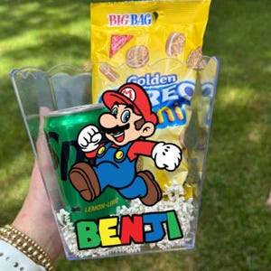 May include: A clear plastic container filled with white paper shred, a green Sprite soda can, and a yellow bag of Golden Oreo Mini cookies. The front of the container features a colorful graphic of Mario and the name Benji in a bold, multi-colored font.