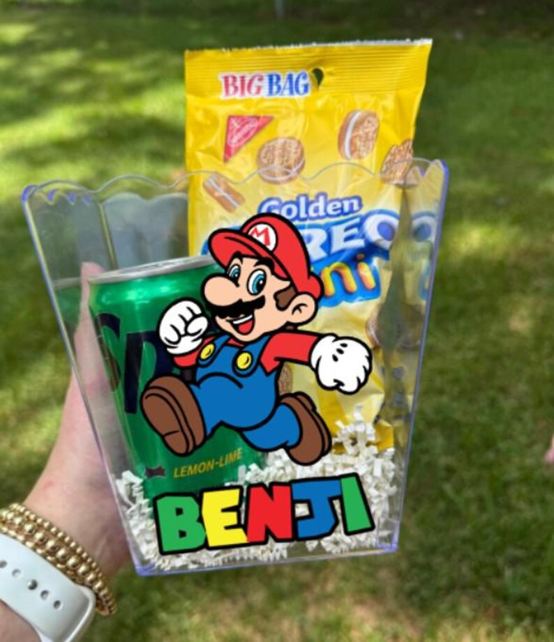 May include: A clear plastic container filled with white paper shred, a green Sprite soda can, and a yellow bag of Golden Oreo Mini cookies. The front of the container features a colorful graphic of Mario and the name Benji in a bold, multi-colored font.