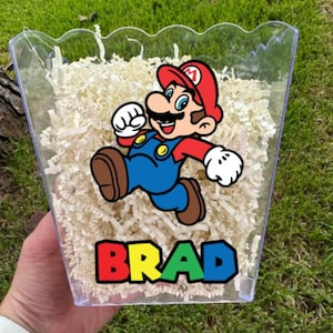 May include: A clear plastic scalloped container filled with shredded white paper filler. The front features a colorful graphic of the character Mario in a red hat, blue overalls, and brown shoes, with the name BRAD printed in bold, multi-colored letters.