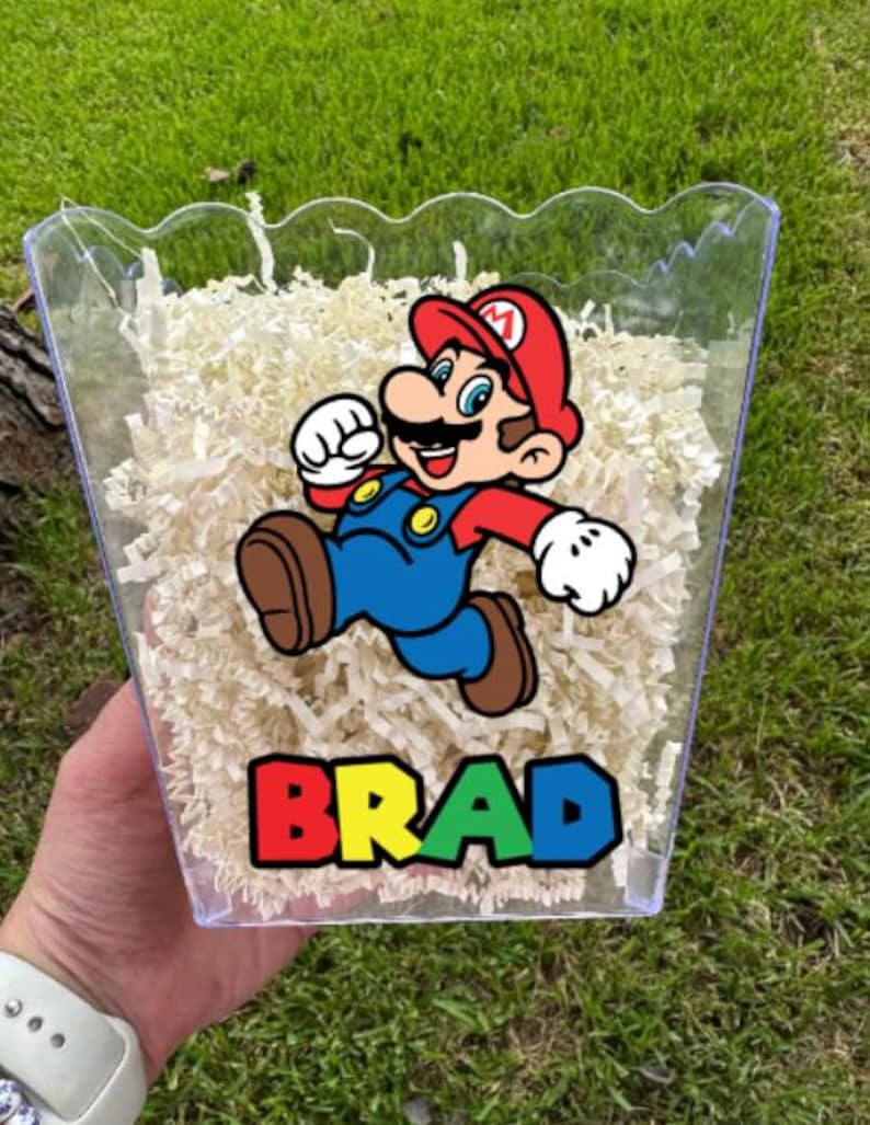 May include: A clear plastic scalloped container filled with shredded white paper filler. The front features a colorful graphic of the character Mario in a red hat, blue overalls, and brown shoes, with the name BRAD printed in bold, multi-colored letters.