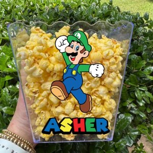 May include: A clear plastic popcorn container filled with yellow popcorn. The container features a graphic of the character Luigi wearing a green hat and blue overalls, with the name ASHER printed in a bold, multi-colored block font below.