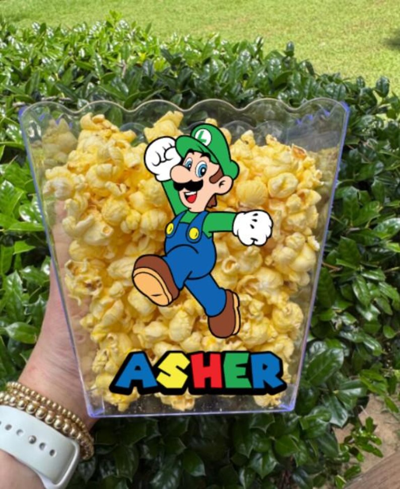 May include: A clear plastic popcorn container filled with yellow popcorn. The container features a graphic of the character Luigi wearing a green hat and blue overalls, with the name ASHER printed in a bold, multi-colored block font below.