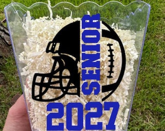 May include: A clear plastic scalloped container filled with light-colored paper shred. The front features a black graphic of a football helmet and ball, with the blue text SENIOR and the blue number 2027 printed below the graphic design.