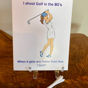 May include: A white card with a cartoon illustration of a woman playing golf. The text on the card reads "I shoot Golf in the 80's When it gets any hotter than that, I Quit!!"