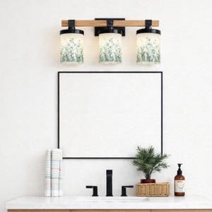 May include: A three-light vanity fixture with a wooden beam and black metal accents. The cylindrical glass shades feature a green and white botanical pattern. Below, a black-framed mirror hangs above a sink with a black faucet and a small potted plant.