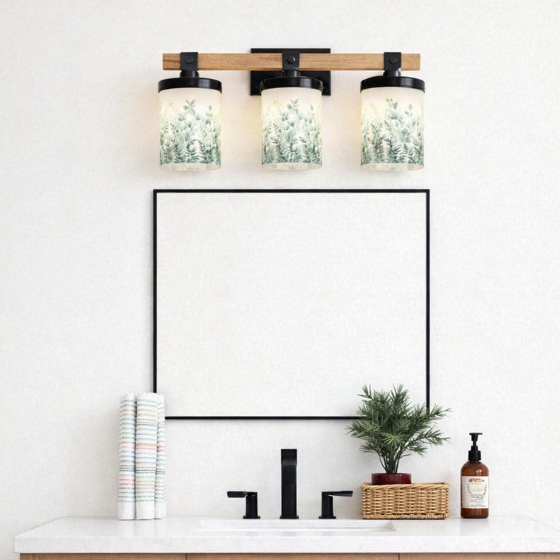 May include: A three-light vanity fixture with a wooden beam and black metal accents. The cylindrical glass shades feature a green and white botanical pattern. Below, a black-framed mirror hangs above a sink with a black faucet and a small potted plant.