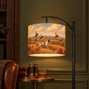 May include: A floor lamp with a black metal stand and a cylindrical lampshade featuring a printed landscape scene. The artwork depicts a white and brown spotted dog running through a golden field with several birds flying in a cloudy sky.
