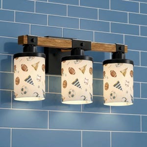 May include: A three-light vanity fixture with a wooden beam and black metal accents. The cylindrical shades feature a sports-themed pattern with footballs, soccer balls, and baseballs. The fixture is mounted on a blue tiled wall.