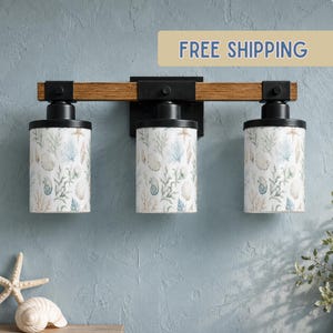 May include: A three-light vanity fixture featuring a horizontal wooden bar with black metal accents and cylindrical glass shades. Each shade displays a printed pattern of seashells, coral, and seaweed in shades of blue, green, and tan.