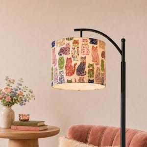 May include: A floor lamp with a black metal curved stand and a cylindrical lampshade featuring a repeating pattern of colorful illustrated cats in various poses and colors, including orange, green, blue, and pink, set against a cream background.