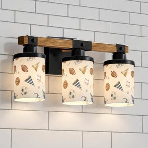 May include: A three-light vanity fixture with a wooden beam and black metal accents. The cylindrical shades feature a sports-themed pattern with footballs, baseballs, soccer balls, and party hats. The fixture is mounted on a white brick wall.