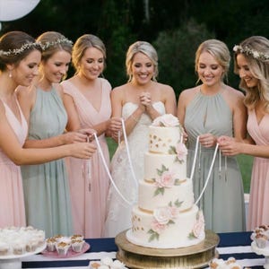 May include: A multi-tiered wedding cake with gold accents and pink floral decorations. The cake is on a gold stand, surrounded by women in pastel dresses holding ribbons. Cupcakes and other desserts are on a table.