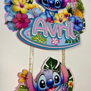 May include: A vibrant cake topper with a cartoon Stitch design, adorned with tropical flowers and the name "Avril". The number "6" is present, indicating a birthday celebration. The design is layered.