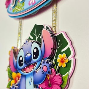 May include: A layered cake topper featuring the character Stitch from Lilo & Stitch. The blue Stitch figure is surrounded by tropical flowers and leaves. The topper also includes the name "Avril" in pink and blue.