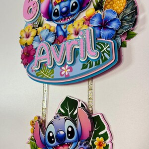 May include: A layered cake topper featuring the character Stitch from Lilo & Stitch. The topper includes the name "Avril" and the number "6", surrounded by tropical flowers, leaves, and pineapples. The design is colourful with pink, blue, yellow, and green accents.