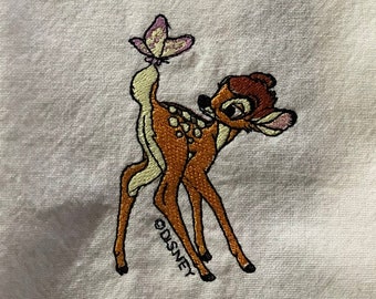 May include: White kitchen towel with an embroidered image of Bambi the deer from Disney. Bambi is brown with white spots and is looking to the right. A purple butterfly is on Bambi's tail. The text "DISNEY" is embroidered below Bambi.