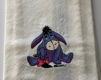 May include: White kitchen towel with a blue and white stripe down the center.  The towel features an embroidered image of Eeyore from Winnie the Pooh sitting down with a red bow on his tail. The embroidery includes the text "Disney".