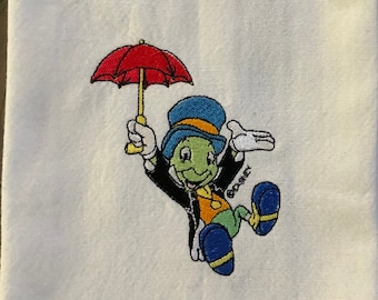 May include: White kitchen towel with an embroidered image of Jiminy Cricket from Disney's Pinocchio. Jiminy is holding a red umbrella and is wearing a green suit, a blue hat, and yellow shoes. The embroidery includes the text "Disney".