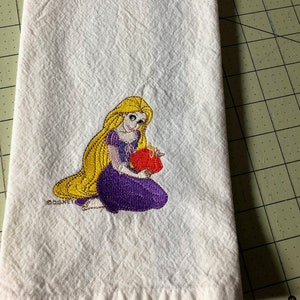 May include: White kitchen towel with an embroidered image of a Disney princess with long blonde hair wearing a purple dress and holding a red heart. The text "DISNEY" is embroidered in the bottom left corner.