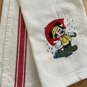 K&ouml;nnte beinhalten: Wei&szlig;es K&uuml;chentuch mit roten Streifen und einem gestickten Bild von Mickey Mouse, der einen Regenschirm im Regen h&auml;lt.