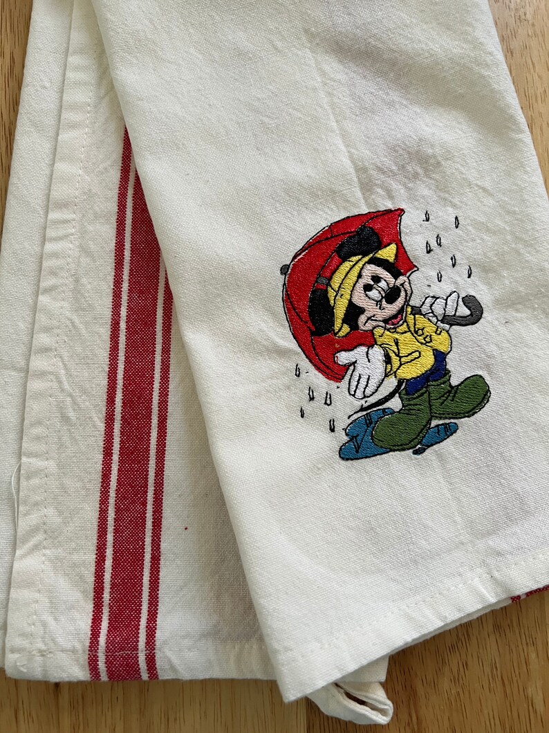 K&ouml;nnte beinhalten: Wei&szlig;es K&uuml;chentuch mit roten Streifen und einem gestickten Bild von Mickey Mouse, der einen Regenschirm im Regen h&auml;lt.