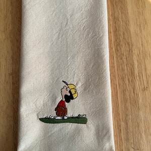 May include: A white kitchen towel with an embroidered image of a cartoon character wearing a baseball cap and holding a baseball glove. The character is looking up and the text "CUFFS" is embroidered below the character.