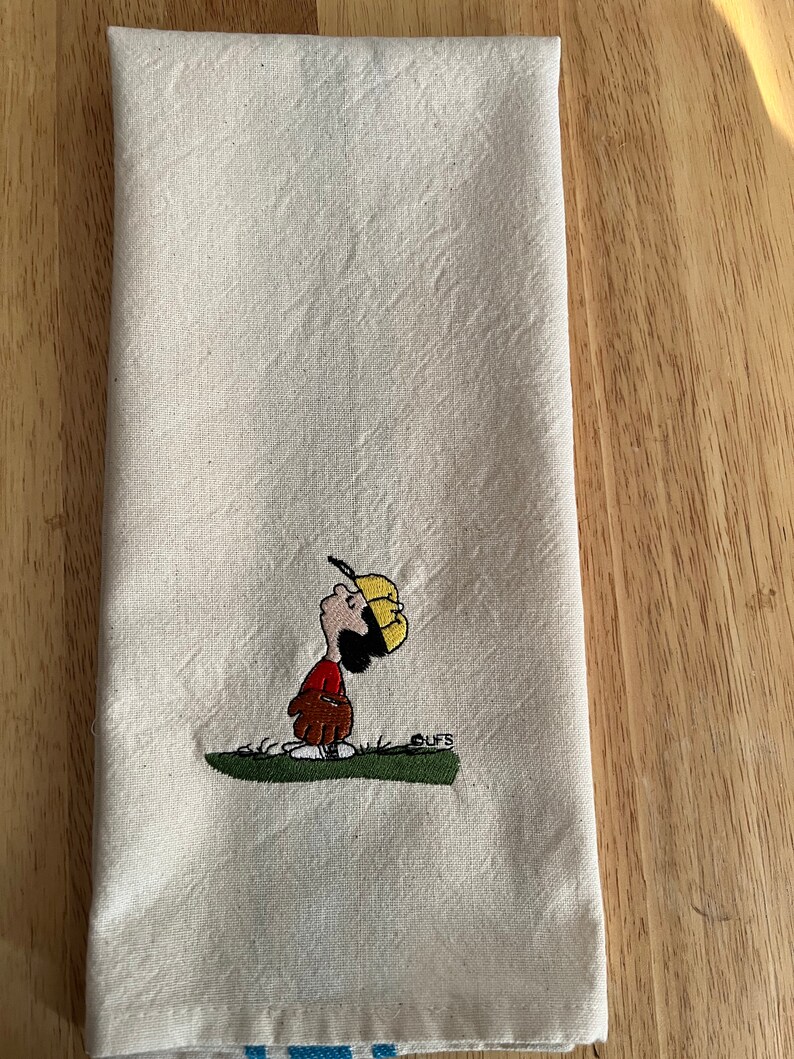 May include: A white kitchen towel with an embroidered image of a cartoon character wearing a baseball cap and holding a baseball glove. The character is looking up and the text "CUFFS" is embroidered below the character.