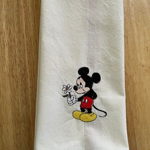 May include: White kitchen towel with an embroidered image of Mickey Mouse holding a white flower. Mickey Mouse is wearing red shorts and yellow shoes. He has a sad expression on his face.