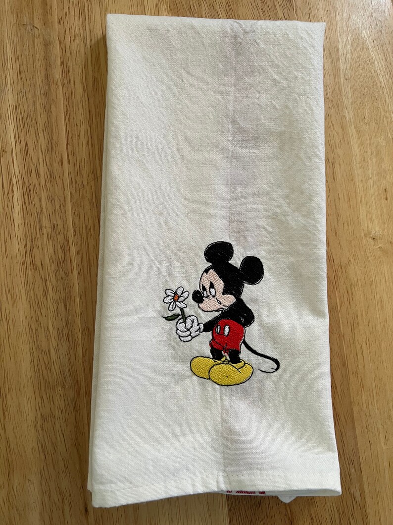 May include: White kitchen towel with an embroidered image of Mickey Mouse holding a white flower. Mickey Mouse is wearing red shorts and yellow shoes. He has a sad expression on his face.