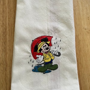 K&ouml;nnte beinhalten: Wei&szlig;es K&uuml;chentuch mit einem gestickten Bild von Mickey Mouse, der einen gelben Regenmantel tr&auml;gt und einen roten Regenschirm h&auml;lt. Um Mickey Mouse herum sind Regentropfen gestickt.