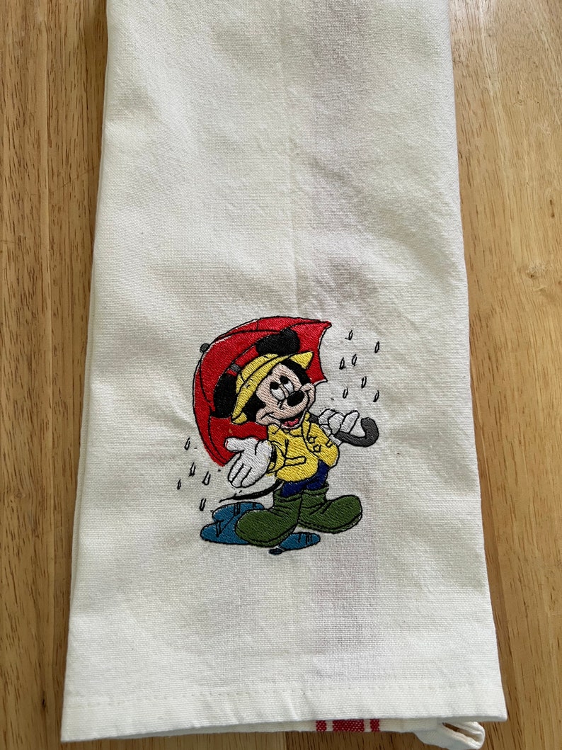 K&ouml;nnte beinhalten: Wei&szlig;es K&uuml;chentuch mit einem gestickten Bild von Mickey Mouse, der einen gelben Regenmantel tr&auml;gt und einen roten Regenschirm h&auml;lt. Um Mickey Mouse herum sind Regentropfen gestickt.
