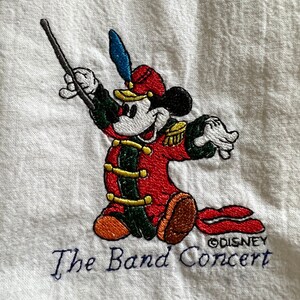 May include: Embroidered image of Mickey Mouse dressed as a band conductor, holding a baton and wearing a red and green uniform. The text "The Band Concert" and "&copy;DISNEY" are embroidered below Mickey.
