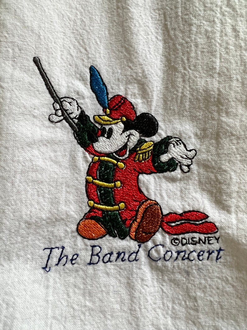 May include: Embroidered image of Mickey Mouse dressed as a band conductor, holding a baton and wearing a red and green uniform. The text "The Band Concert" and "&copy;DISNEY" are embroidered below Mickey.