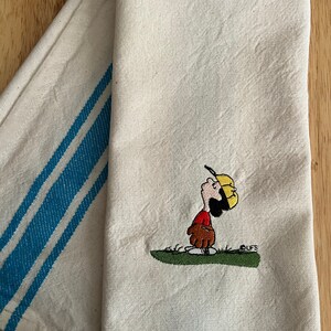 May include: A white tea towel with blue stripes on one side and an embroidered image of a cartoon character playing baseball on the other side. The character is wearing a red shirt, yellow hat, and brown glove. The text "CUFS" is embroidered below the character.