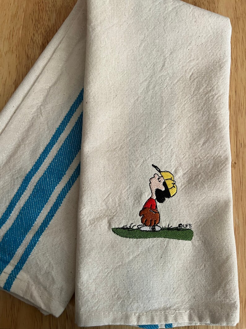 May include: A white tea towel with blue stripes on one side and an embroidered image of a cartoon character playing baseball on the other side. The character is wearing a red shirt, yellow hat, and brown glove. The text "CUFS" is embroidered below the character.