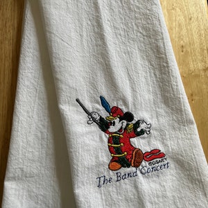 May include: White kitchen towel featuring an embroidered image of Mickey Mouse dressed as a band conductor. The text "The Band Concert" and "&copy;DISNEY" are also embroidered on the towel.