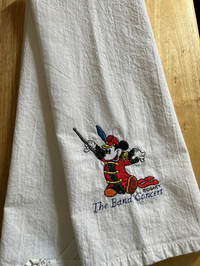 May include: White kitchen towel featuring an embroidered image of Mickey Mouse dressed as a band conductor. The text "The Band Concert" and "&copy;DISNEY" are also embroidered on the towel.