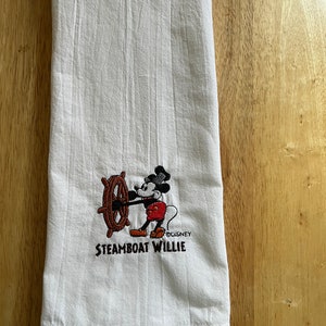K&ouml;nnte beinhalten: Wei&szlig;es K&uuml;chentuch mit einem gestickten Bild von Mickey Mouse als Steamboat Willie. Das Bild zeigt Mickey Mouse, der ein Schiffsrad h&auml;lt und ein rotes Hemd und schwarze Shorts tr&auml;gt. Der Text "Steamboat Willie" ist unter dem Bild gestickt.