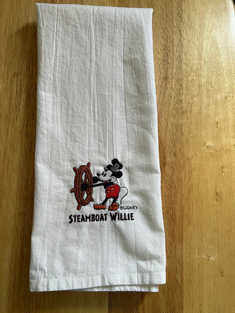 K&ouml;nnte beinhalten: Wei&szlig;es K&uuml;chentuch mit einem gestickten Bild von Mickey Mouse als Steamboat Willie. Das Bild zeigt Mickey Mouse, der ein Schiffsrad h&auml;lt und ein rotes Hemd und schwarze Shorts tr&auml;gt. Der Text "Steamboat Willie" ist unter dem Bild gestickt.
