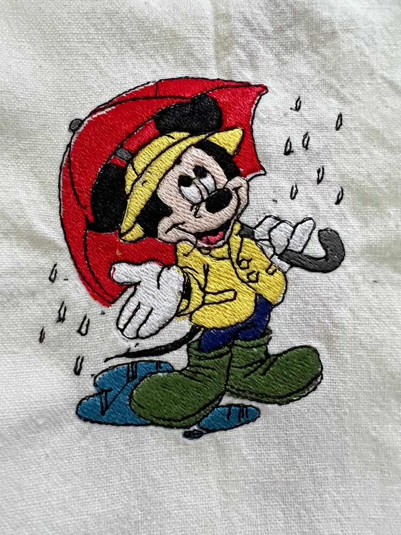 K&ouml;nnte beinhalten: Gesticktes Bild von Mickey Mouse, der einen gelben Regenmantel tr&auml;gt und einen roten Regenschirm h&auml;lt. Er steht in einer Pf&uuml;tze mit Regentropfen, die um ihn herum fallen.