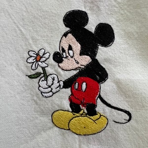 May include: Embroidered image of Mickey Mouse holding a white daisy, with a sad expression on his face. He is wearing red shorts and yellow shoes.