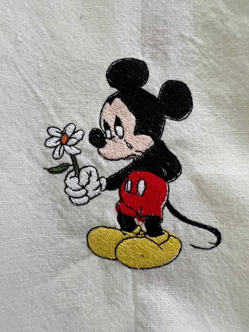 May include: Embroidered image of Mickey Mouse holding a white daisy, with a sad expression on his face. He is wearing red shorts and yellow shoes.