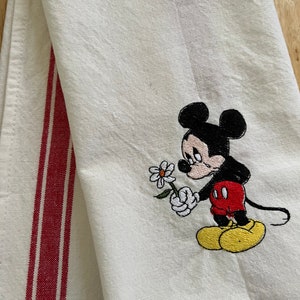 May include: A white kitchen towel with red stripes on the side. The towel features an embroidered image of Mickey Mouse holding a flower.