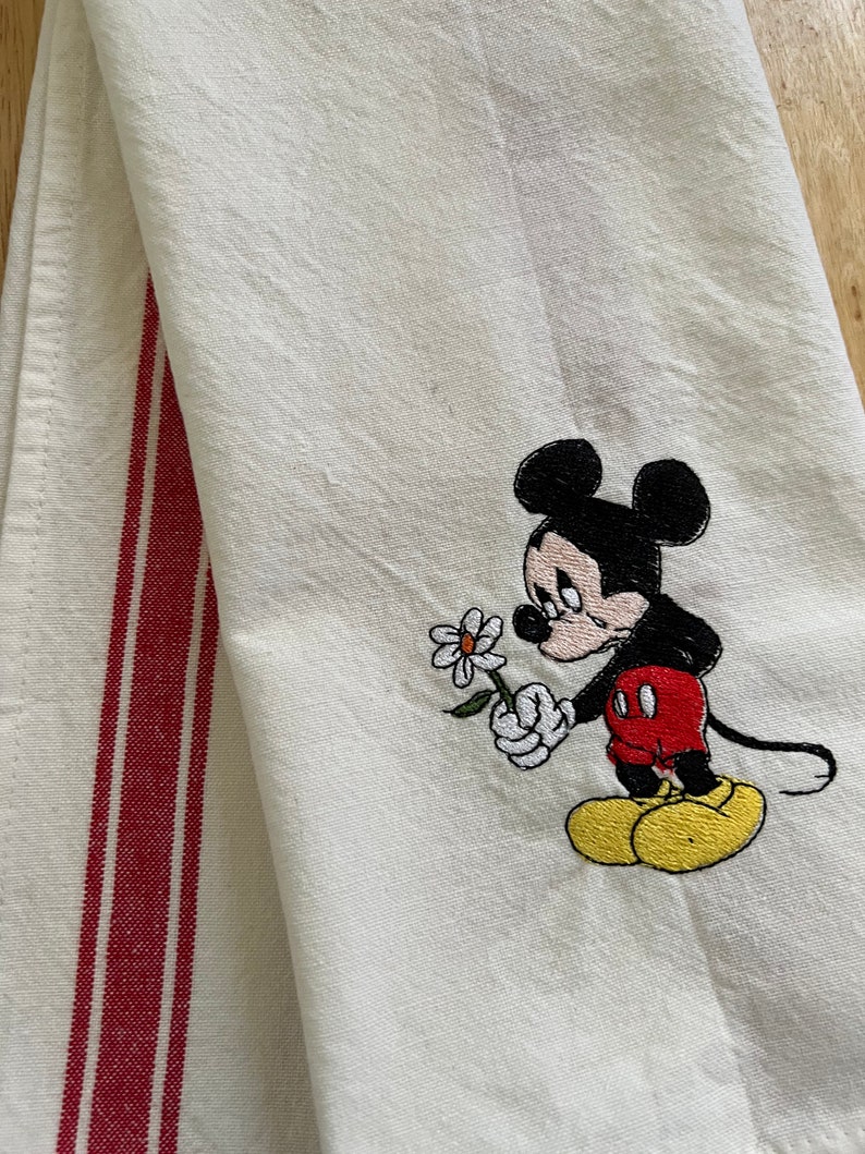 May include: A white kitchen towel with red stripes on the side. The towel features an embroidered image of Mickey Mouse holding a flower.