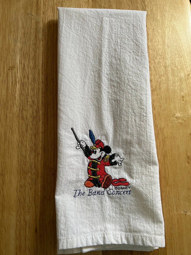 May include: White kitchen towel featuring an embroidered image of Mickey Mouse dressed as a band conductor. The text "The Band Concert" and "&copy;DISNEY" are also embroidered on the towel.