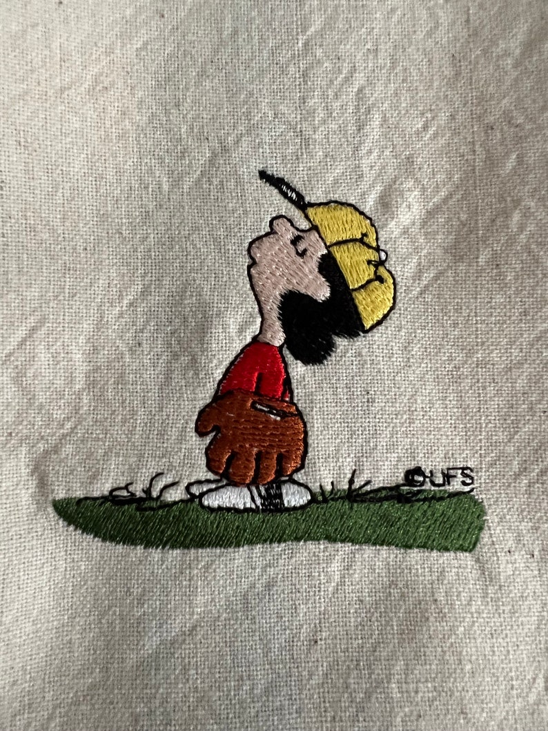 May include: Embroidered image of a cartoon character wearing a yellow baseball cap and a red shirt, holding a brown baseball glove. The character is standing on green grass with the word "CUFS" embroidered in white below.