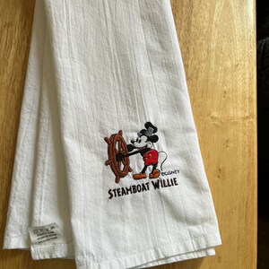 K&ouml;nnte beinhalten: Wei&szlig;es K&uuml;chentuch mit einem gestickten Bild von Mickey Mouse als Steamboat Willie. Das Bild zeigt Mickey Mouse, der das Steuerrad eines Schiffes h&auml;lt. Der Text "Steamboat Willie" und "&copy;Disney" sind unter dem Bild gestickt.