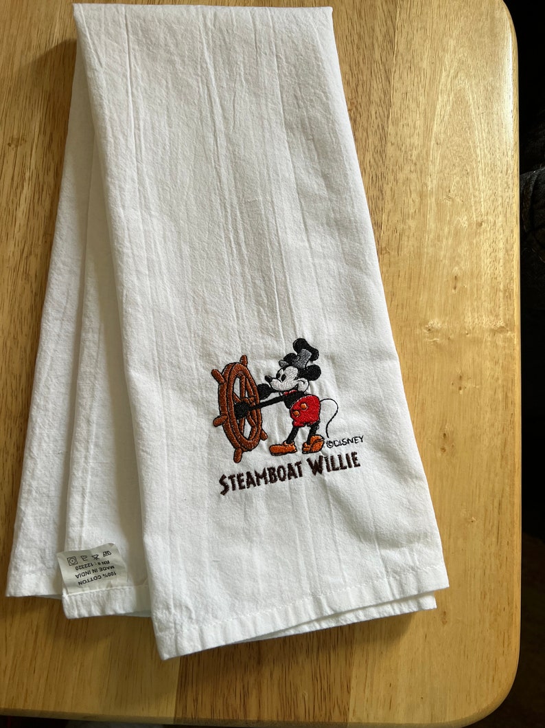 K&ouml;nnte beinhalten: Wei&szlig;es K&uuml;chentuch mit einem gestickten Bild von Mickey Mouse als Steamboat Willie. Das Bild zeigt Mickey Mouse, der das Steuerrad eines Schiffes h&auml;lt. Der Text "Steamboat Willie" und "&copy;Disney" sind unter dem Bild gestickt.