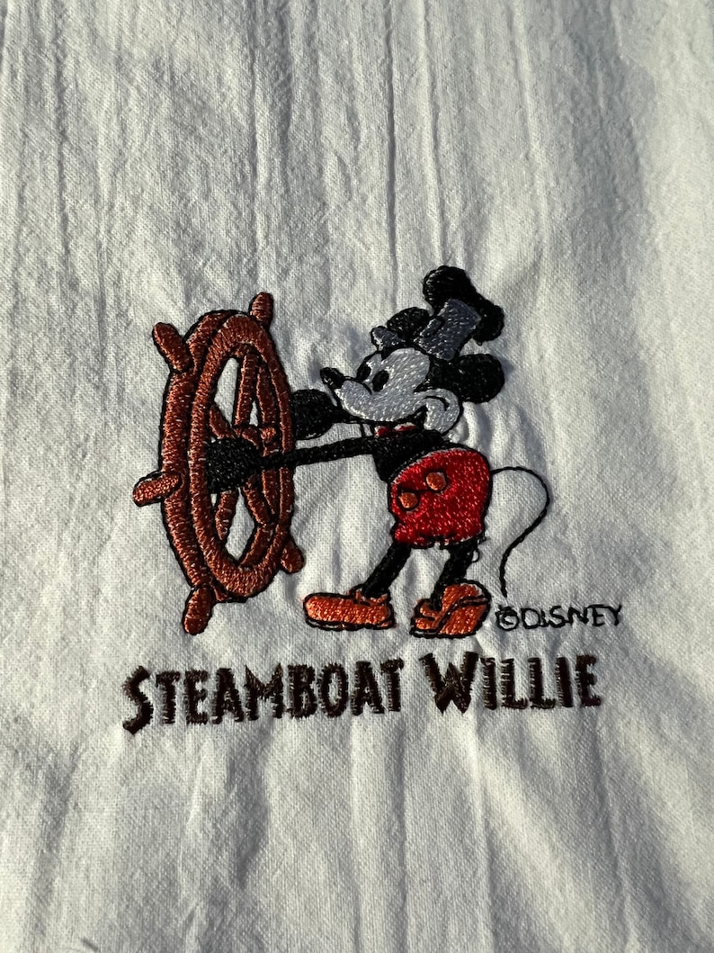 K&ouml;nnte beinhalten: Wei&szlig;es besticktes Hemd mit einem Cartoon-Bild von Mickey Mouse als Steamboat Willie. Mickey h&auml;lt ein braunes Schiffsrad und tr&auml;gt ein rot-schwarzes Outfit. Der Text "Steamboat Willie" und "&copy;Disney" ist unter dem Bild gestickt.